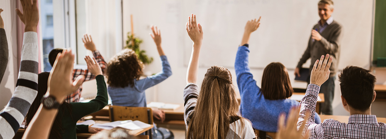 Student in a classroom raise their hands, and their teacher points to one of them.