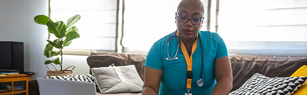 Woman wearing medical scrubs, a lanyard with ID card, and a stethoscope around her neck sitting on a couch looking at a book with an open laptop nearby.