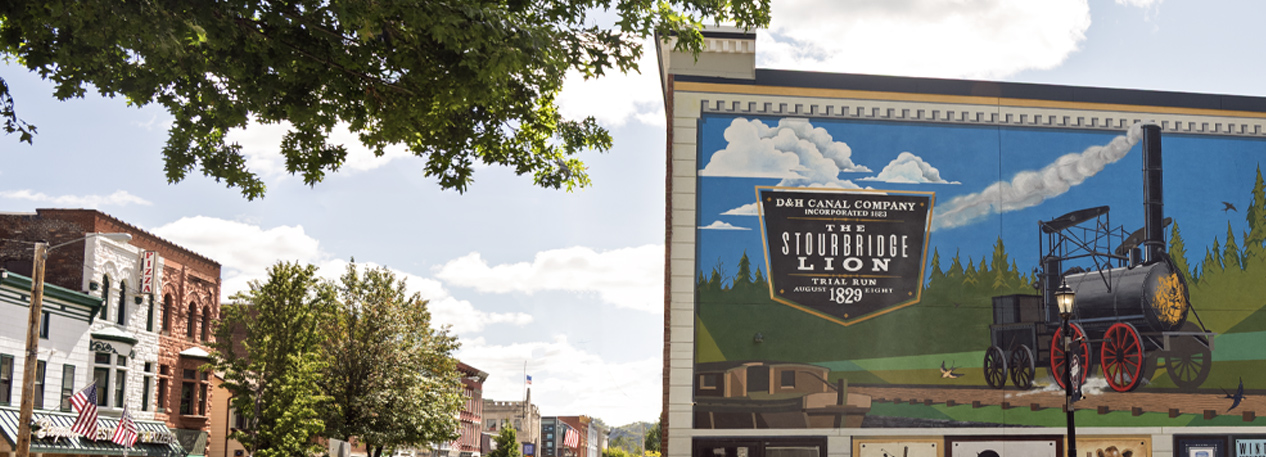A mural on the side of a building in Honesdale, Pennsylvania depicts a old fashioned locomotive train. Leafy tree branches are in the foreground and a main street's building sit in the background of the shot.
