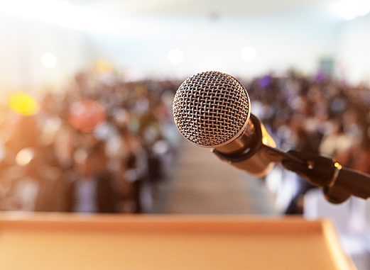 Close up of a microphone at a podium with an audience in the background.