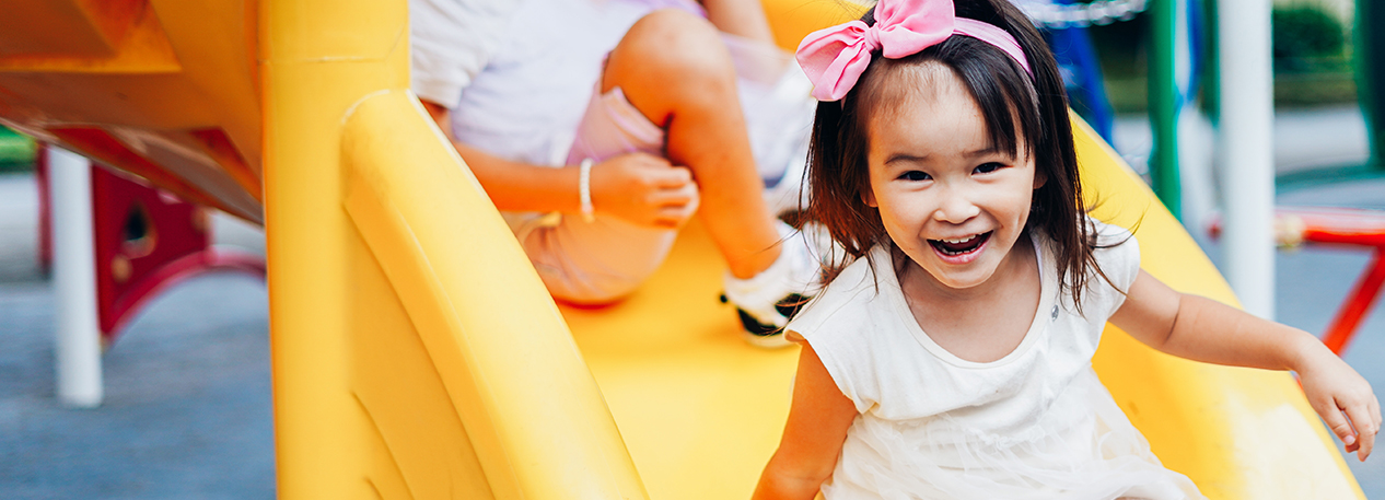 A smiling girl wearing a pink bow slides down a yellow slide.