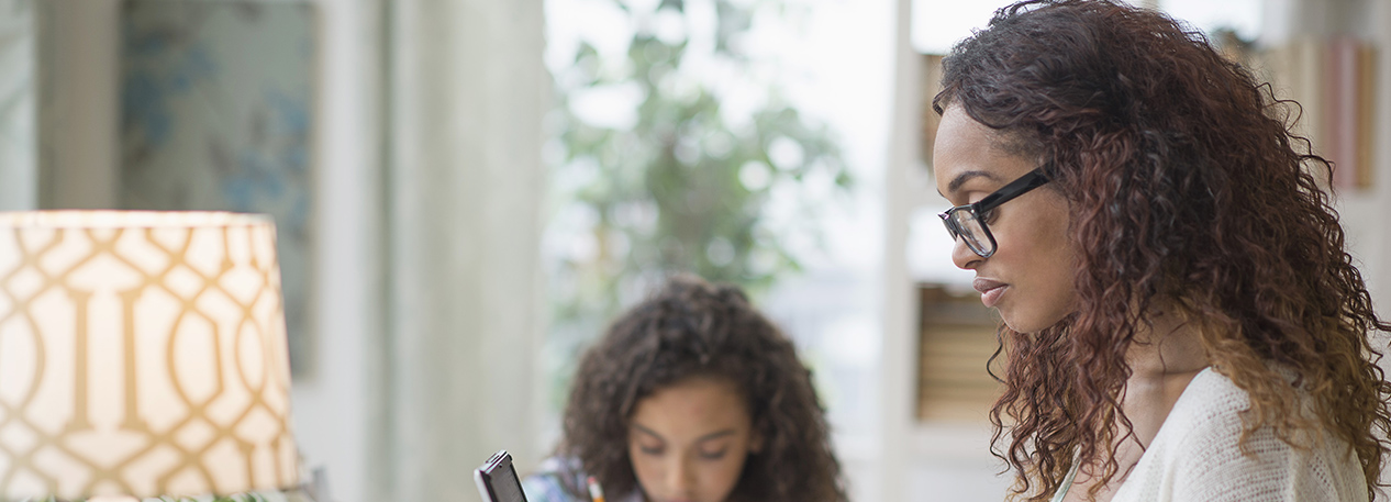Woman working at a laptop with her daughter writing in the background.