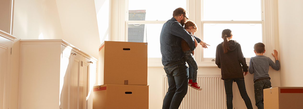 Man and three children looking out of a window in a room with moving boxes stacked on the floor.