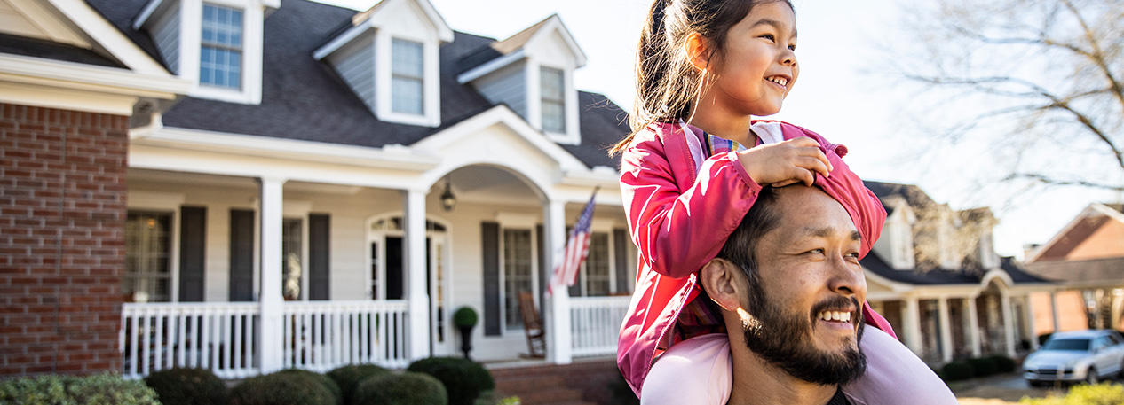 A father stands in front of his home with his daughter sitting on his shoulders.