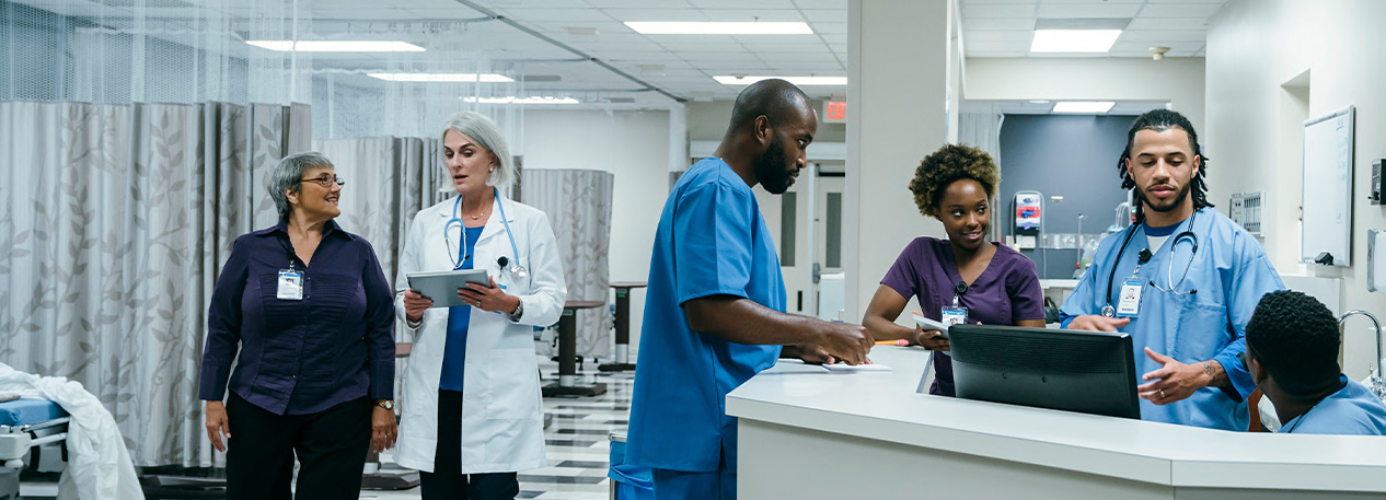 A view of a hospital floor. Nurses meet at a desk and a doctor converses with another employee as they walk down the hall.