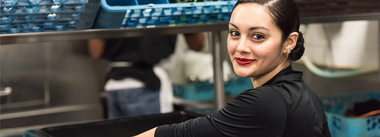 A Hispanic woman looks over her should and smiles as she works in a kitchen.