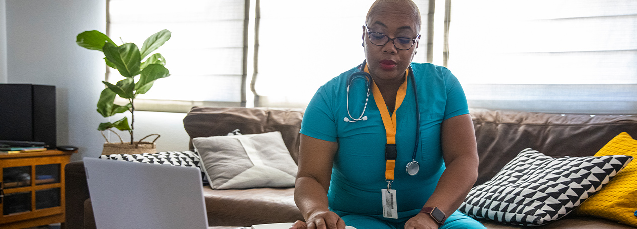 Woman wearing medical scrubs, a lanyard with ID card, and a stethoscope around her neck sitting on a couch looking at a book with an open laptop nearby.