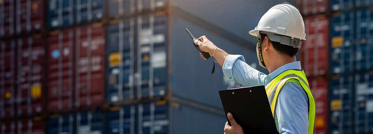 Worker in hardhat, holding clipboard and pointing a walkie-talkie at shipping containers