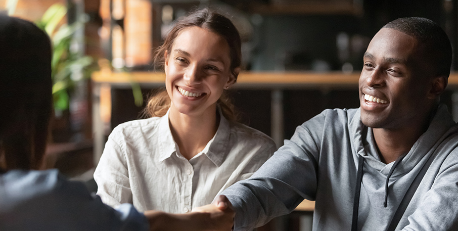 A happy man reaches out to shake a person's hand across a desk. A happy woman looks on.