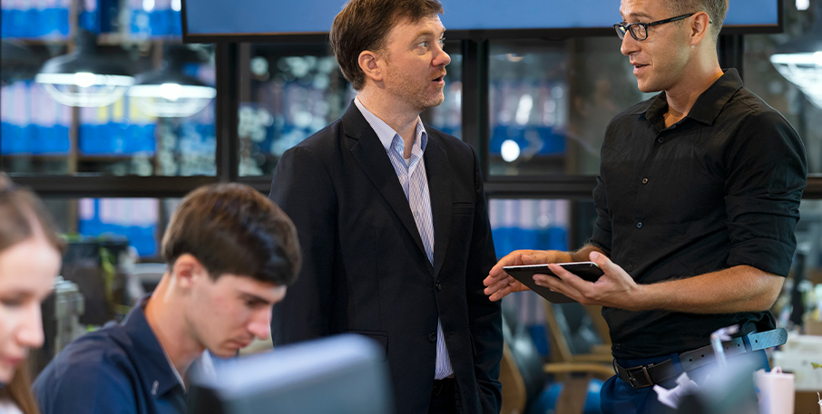 Two coworkers talk while two others type at computers in a modern office setting.