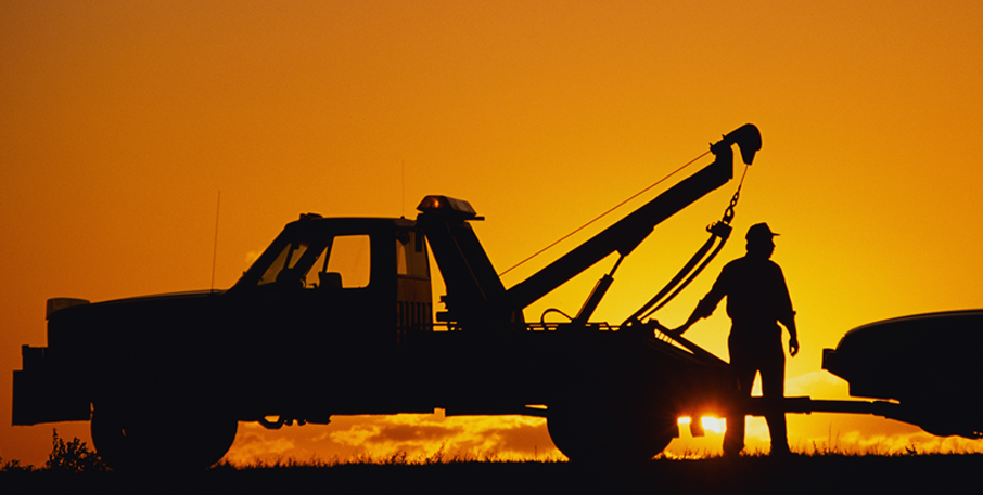 Backlit by an orange sunset, a man connects a car to a tow truck.