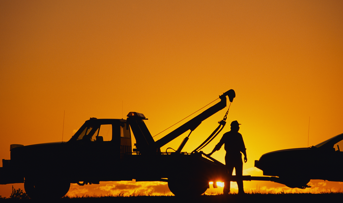 Backlit by an orange sunset, a man connects a car to a tow truck.