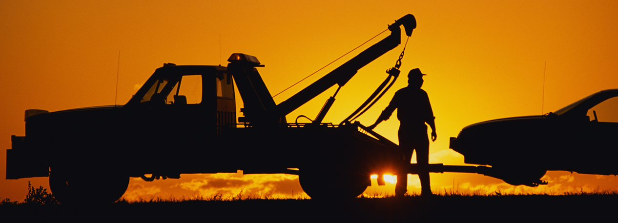 Backlit by an orange sunset, a man connects a car to a tow truck.