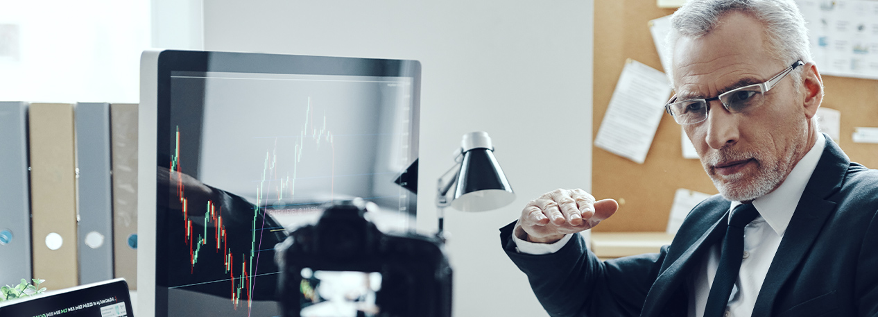 Man wearing a suit and glasses, sitting at a desk recording a video on a camera mounted on a tripod. The desk has a large computer monitor and an open laptop.