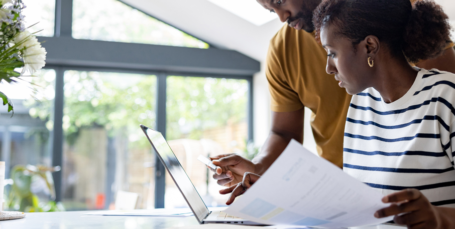A couple reviews information on a laptop and a printed document together.