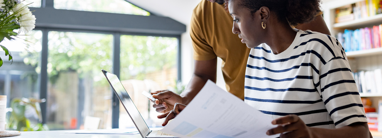 A couple reviews information on a laptop and a printed document together.