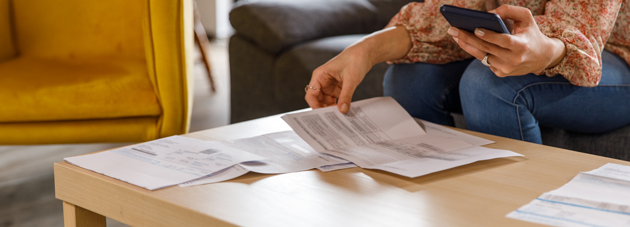 A woman sits on a couch and reviews documents spread across a coffee table. She reviews a message on her cell phone, as well.