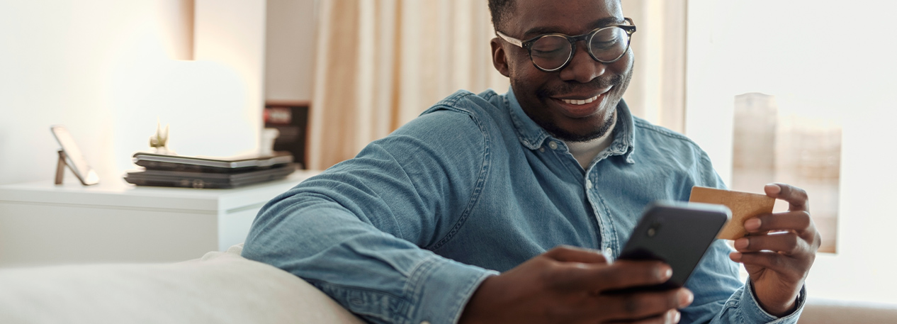 Man sitting on a sofa looking at a cell phone in one hand and holding a credit card in another.