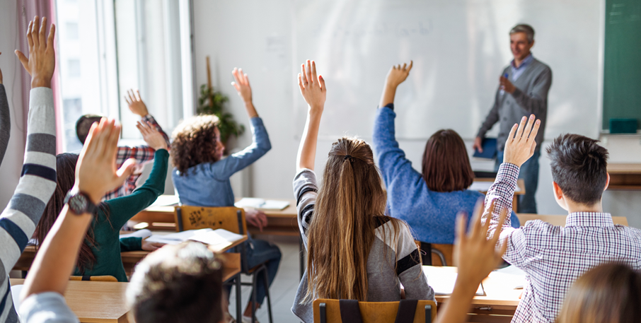 Students raise their hands in a classroom, while a teacher decides who to call on.