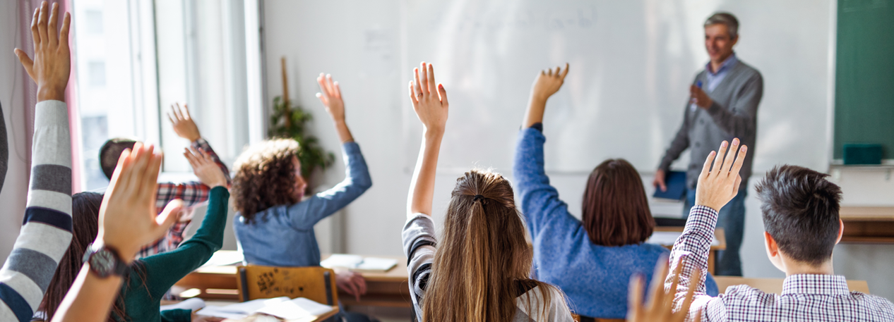 Students raise their hands in a classroom, while a teacher decides who to call on.