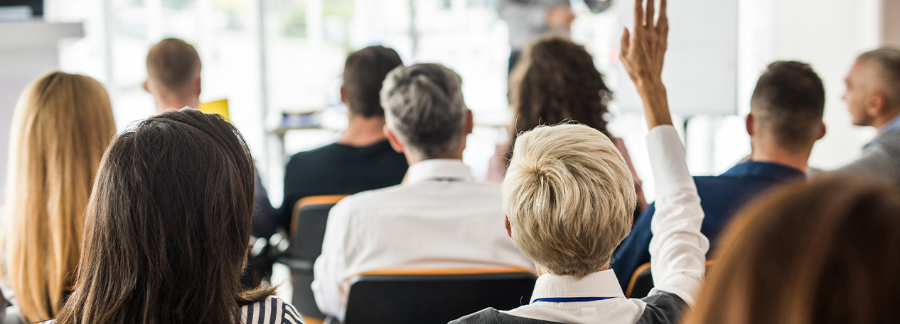 Viewed from the back, an audience looks toward the presenter, and one member of the audience has their hand raised.