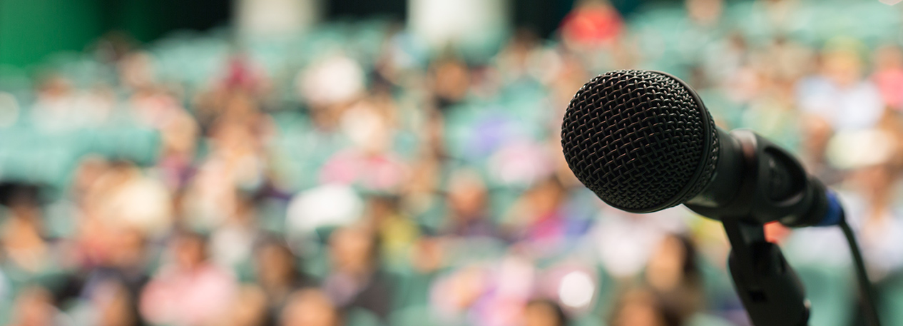 Close-up of a microphone in a stand with an audience sitting in seats in the background.