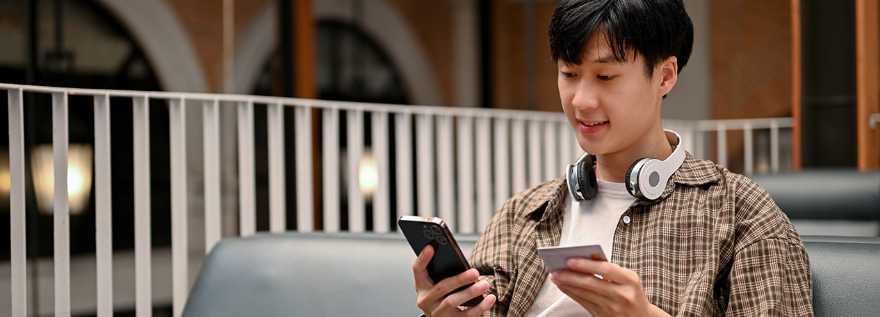 A college student wears headphones around his neck, and he holds a credit in one hand and a cell phone in the other.