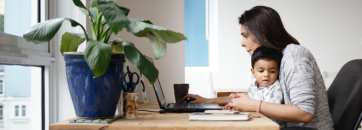 Woman working at home, sitting at a table with a laptop computer, holding a young child on her lap.