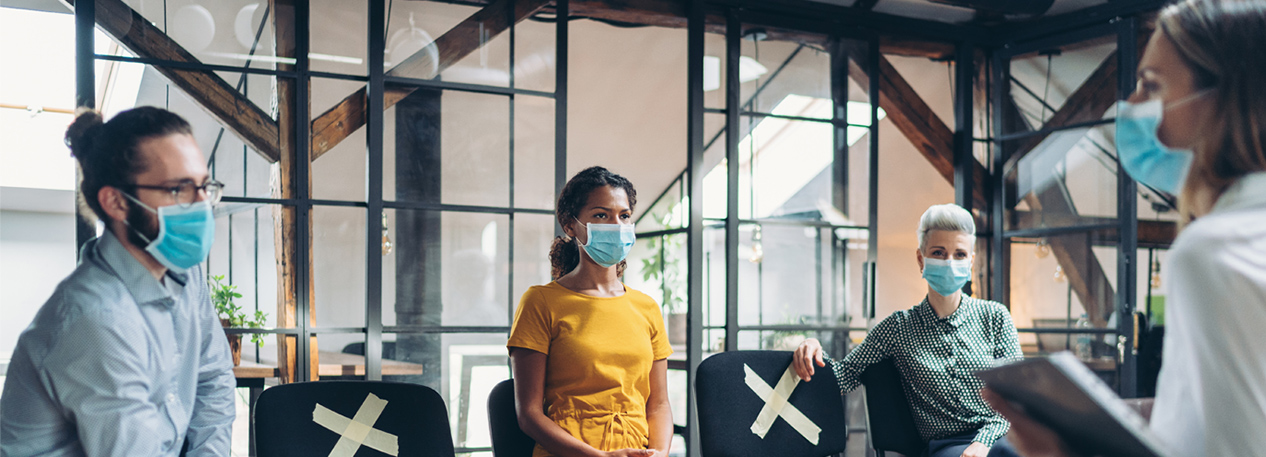 Coworkers with masks in an office, sitting socially distant, with an "x" marking seats not to be used between them.