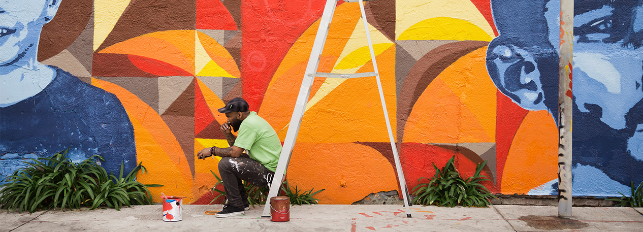 Painter resting on a ladder in front of a brightly colored wall mural featuring two children’s faces