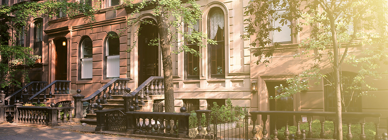 Front entrances of three brownstone townhouses on a tree-lined street