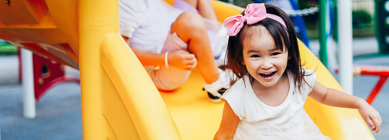 Young girl in a pink bow hairband smiling as she goes down a yellow spiral slide at a playground.