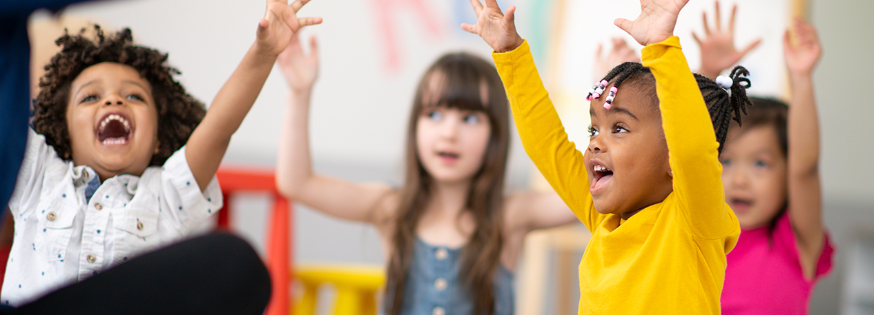 Four children in a classroom raising their arms with excitement and bright smiles across their faces.