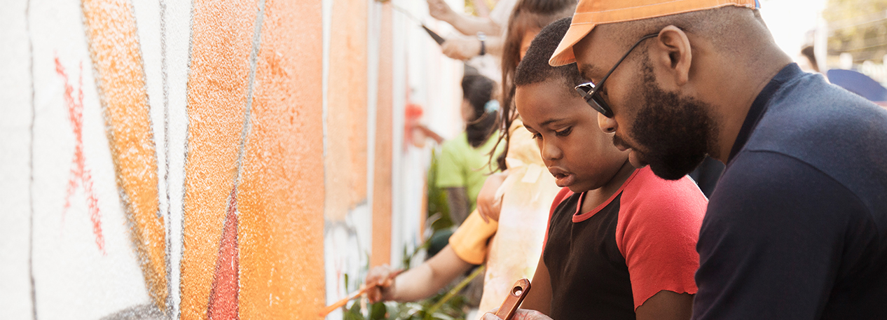 A man dips a brush in orange paint while a child watches. They  are painting a mural.