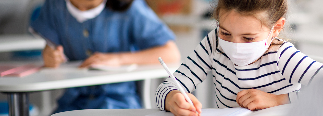 Young children wearing masks, sitting at school desks and writing.