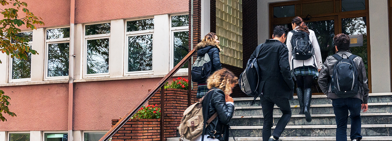 Students in coats and backpacks walking up the steps outside of a school.