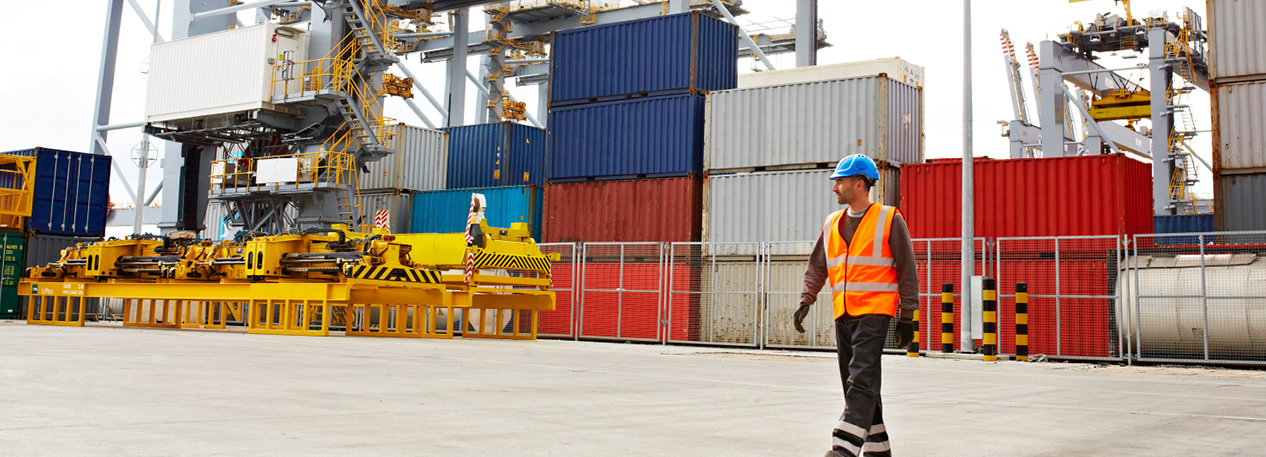Man in a hardhat and bright orange vest walking in front of a stack of shipping containers