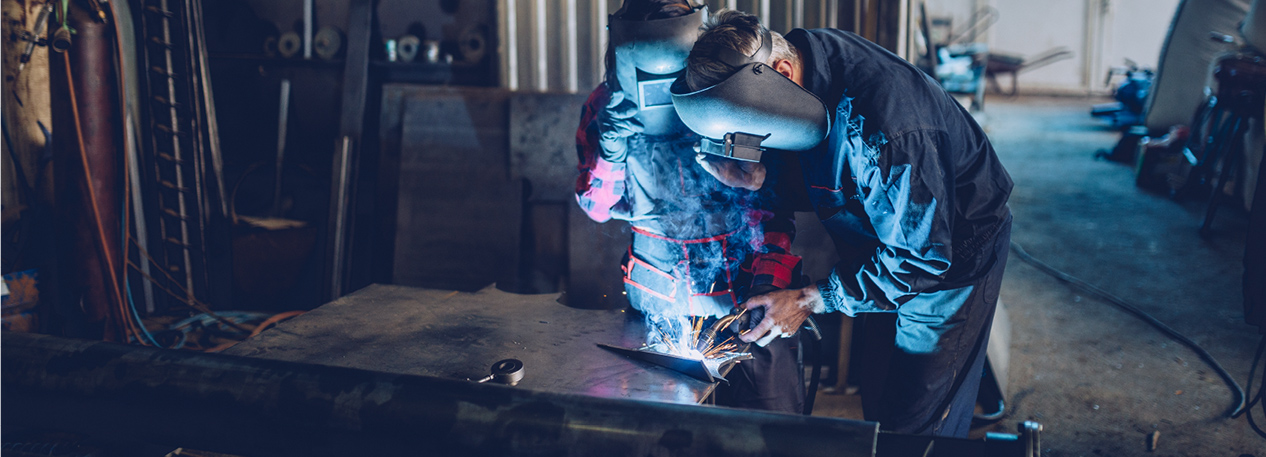 Two workers in a shop with masks and aprons welding a piece of metal