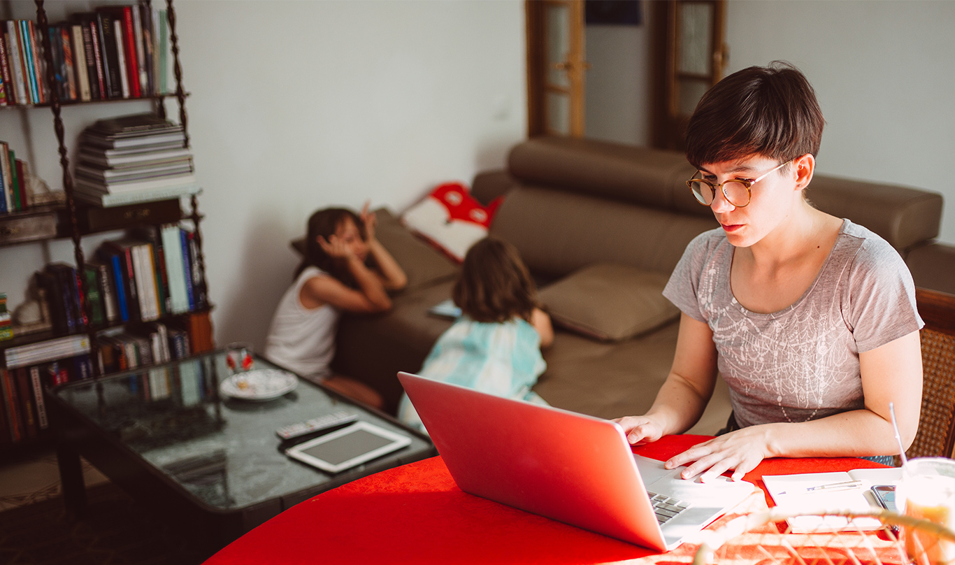 A mother works on a laptop while her two children play in the background.