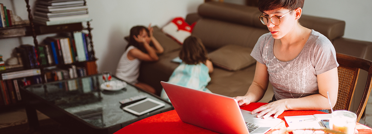 A mother works on a laptop while her two children play in the background.