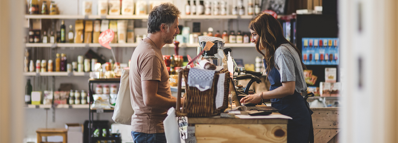 Man with shopping bag being checked out at a register by a woman in a blue apron in a gourmet food store