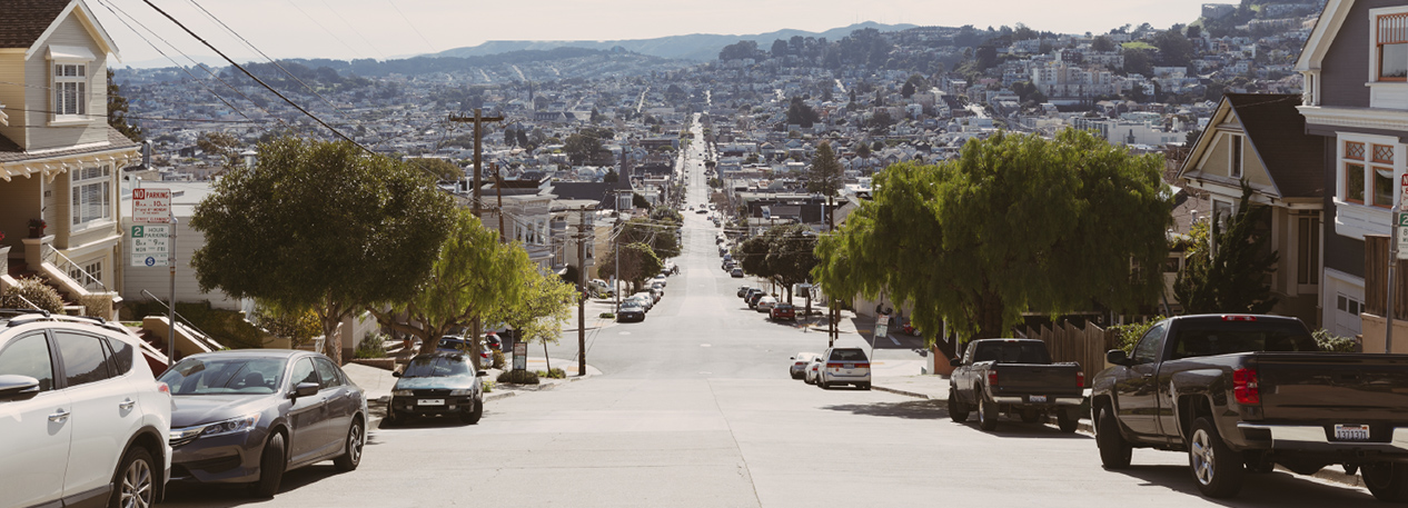 Long, straight road, disappearing into the distance in a hilly city, lined by residential houses and parked cars