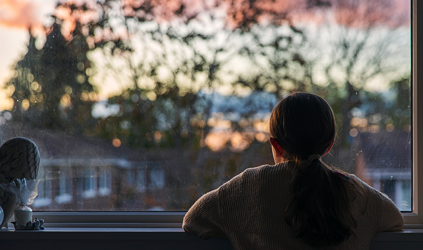 Silhouette of a child looking out a window at a sunset.