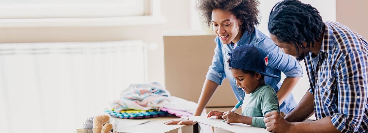 A mom and dad help their school-aged son label the contents of the moving boxes that surround them.