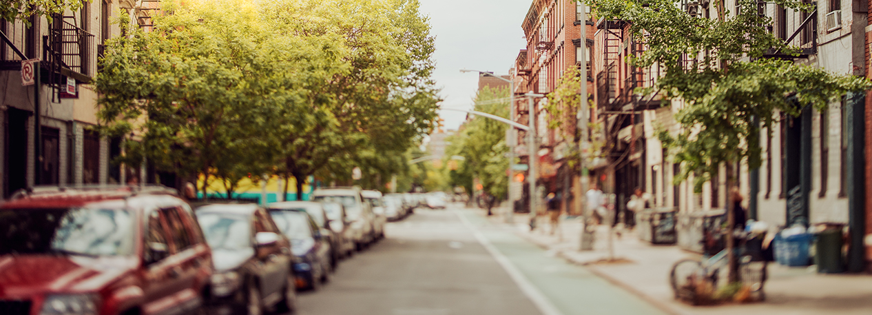 A city street lined with trees, showing residential buildings and cars parked on one side of the road.