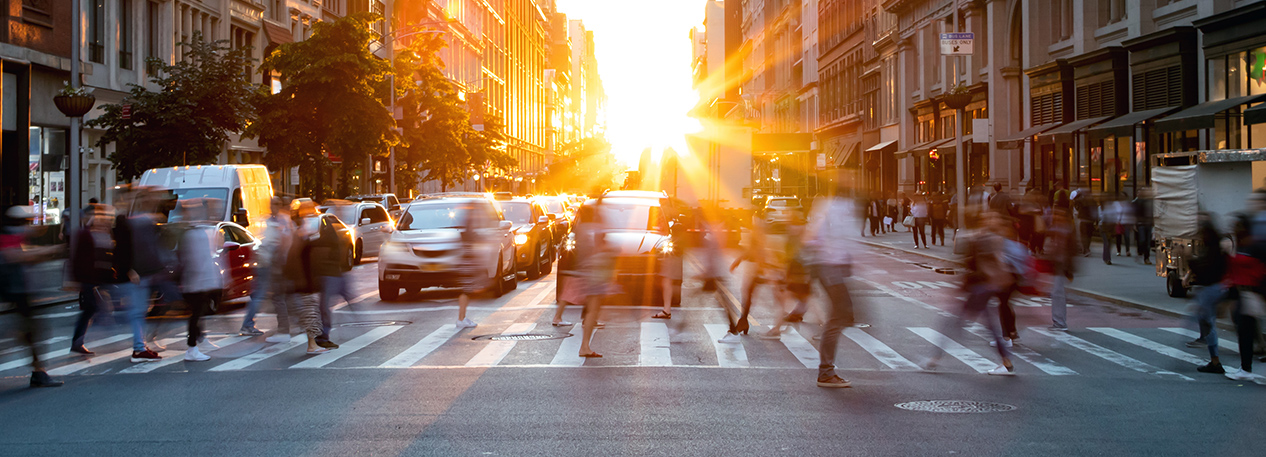 People walking across a city crosswalk with the sun setting in the background.