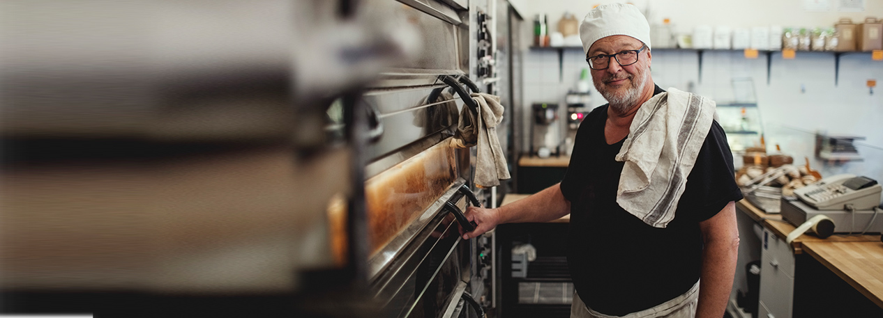 Man in a bakery with a hat and towel over his shoulder, holding the handle of the oven door