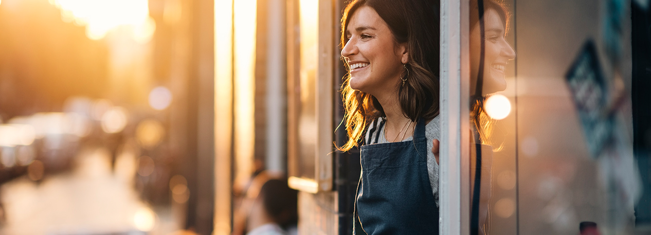 Woman in green apron looking out of storefront door, smiling