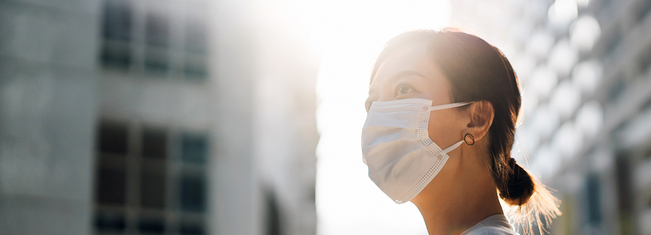 The sun shines through city builds on a woman wearing a mask.