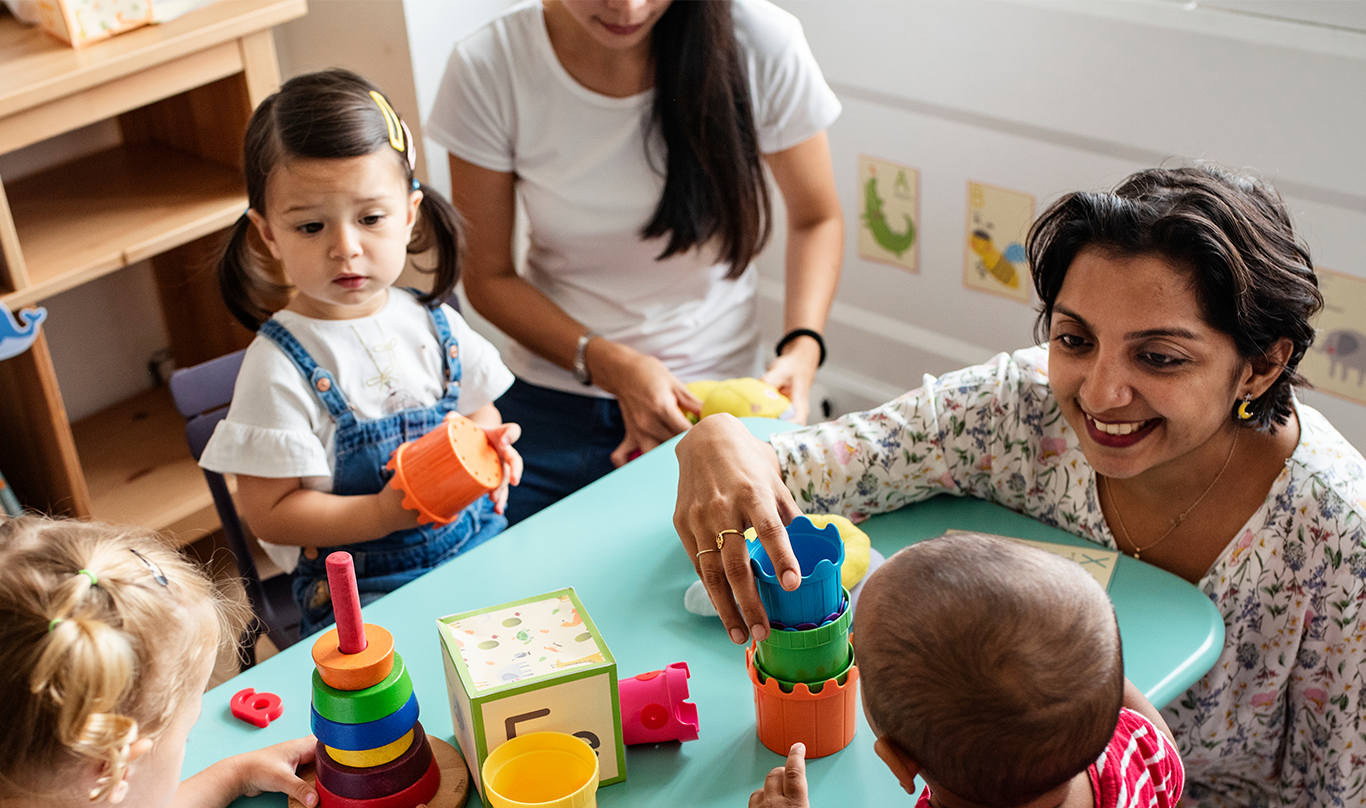 Young children at a daycare center play with toys and interact with their teacher.